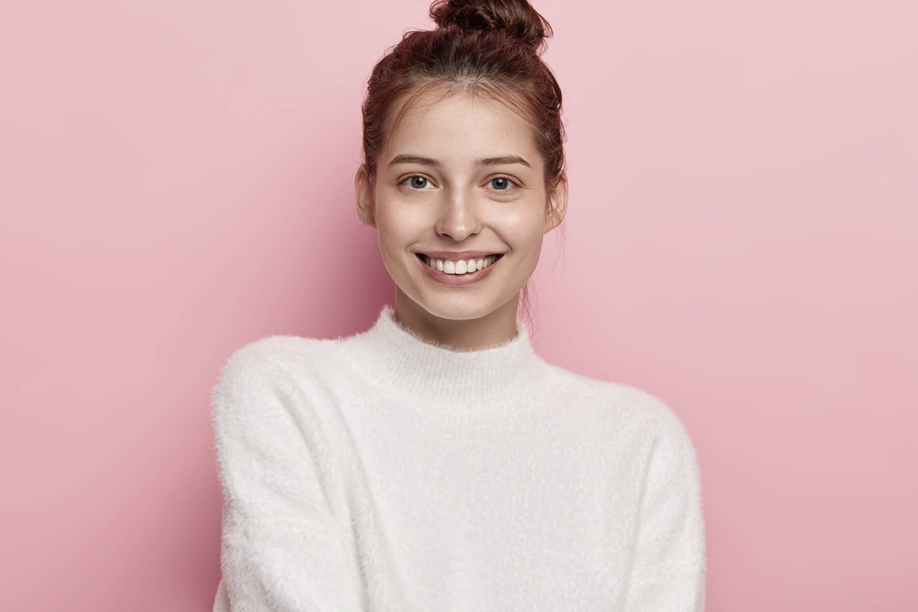 tender-feminine-woman-with-blue-eyes-smiles-pleasantly-has-toothy-smile-wears-white-comfortable-sweater-looks-directly-camera-isolated-pink-background